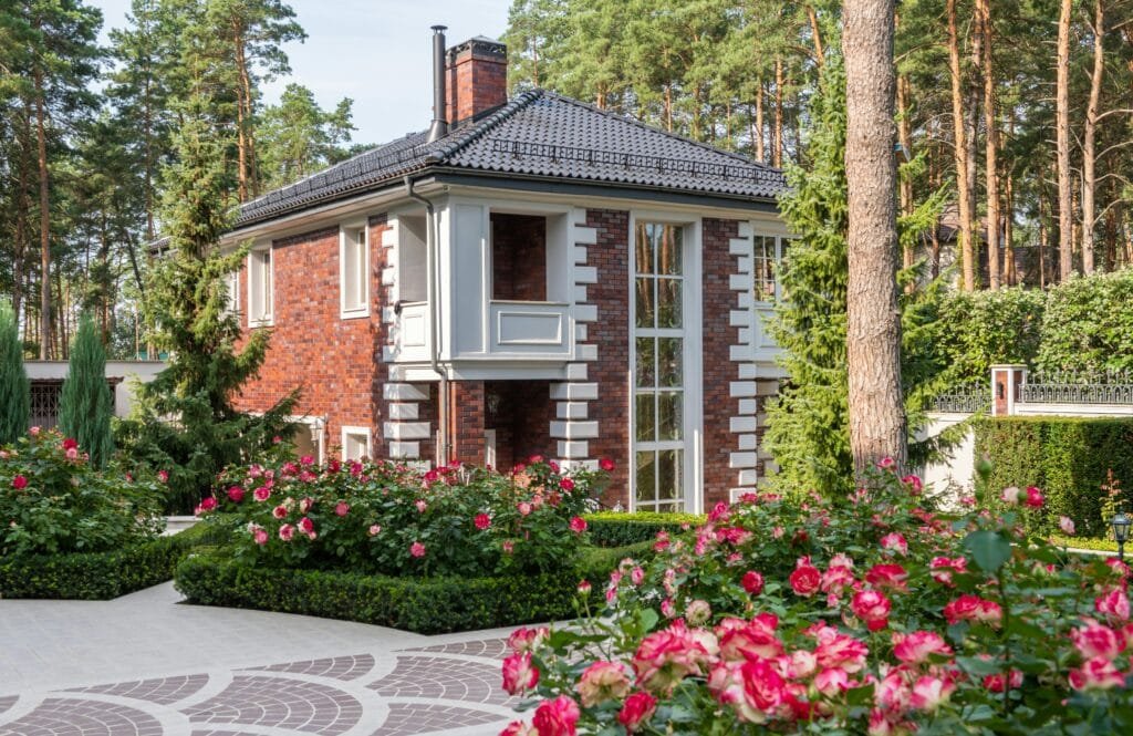 Flowers and green plants with trees in yard with pathway near cottage exterior in sunny summer day